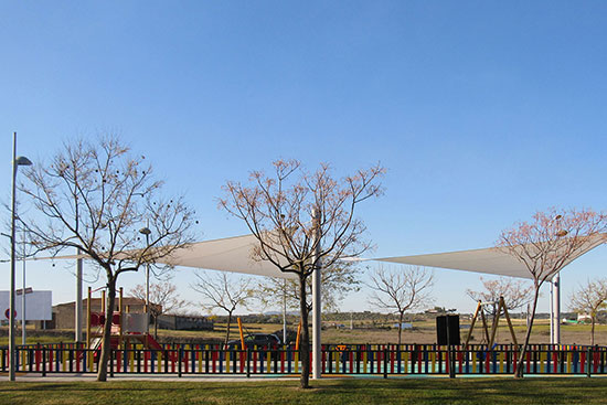 A playground with colourful fencing, trees and tension sail shade structures over the play area.